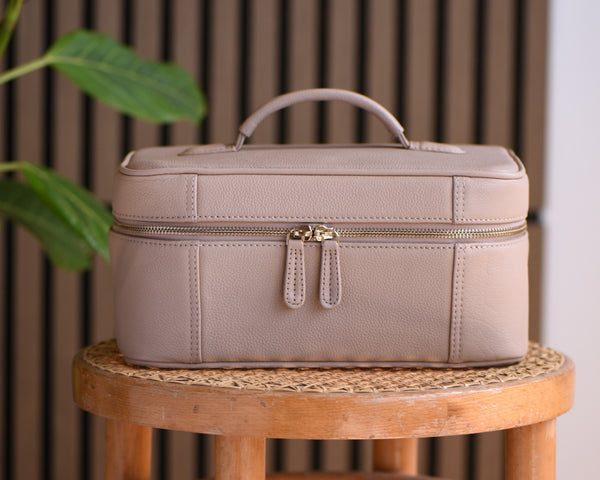A structured light beige leather cosmetic case with a top handle and double zipper closure, displayed on a wooden stool against a brown slatted background with a green plant leaf on the left.