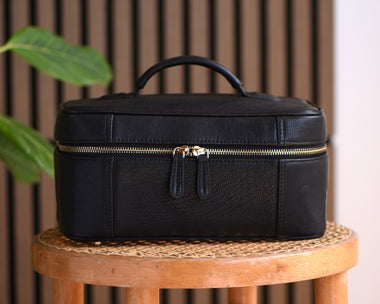 A black leather cosmetic case with a top handle and gold-tone zipper, displayed on a wooden stool against a brown slatted wall with green leaves entering from the left.