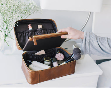 A brown full grain leather makeup bag with a chocolate brown interior, shown open on a white bedside table with a hand lifting the lid. Cosmetics and brushes are visible inside, with a vase of baby's breath and a white lamp in the background.
