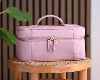 A light pink full grain leather makeup case with a top handle and zipper closure, displayed on a wooden stool against a brown slatted background with a green plant leaf on the left.