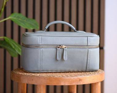 A light green leather cosmetic bag with a top handle and gold zipper, sitting on a wooden stool in front of a brown slatted wall with a green plant on the left.