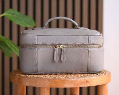 A light gray leather cosmetic case with a top handle and gold zipper, displayed on a wooden stool next to a green plant against a slatted wood and white wall background.