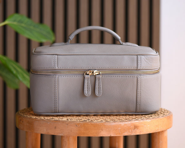 A light gray leather cosmetic case with a top handle and gold zipper, displayed on a wooden stool next to a green plant against a slatted wood and white wall background.