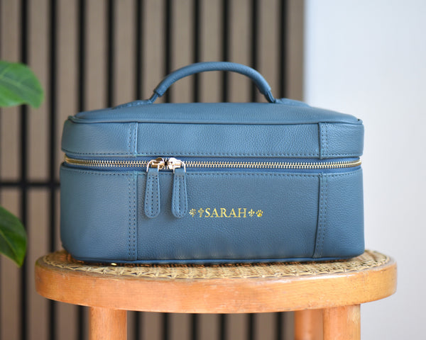A teal pebbled leather cosmetic bag with a top handle and gold zipper, personalized with 'SARAH' in gold lettering, sitting on a wooden stool.