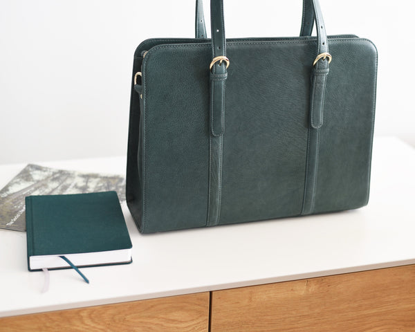 A green leather laptop bag with a sleek design placed on a white surface next to a closed notebook and a smartphone.