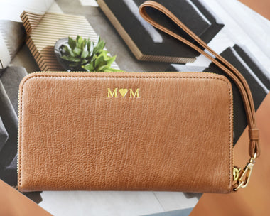A brown leather ziparound wallet with a detachable wrist strap, embossed with the initial 'M' on the front, displayed on a flat surface with books and a plant in the background.