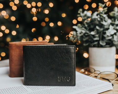 A black leather wallet with a hot stamped monogram on the front, placed on a table with a blurred background featuring Christmas lights and a plant.