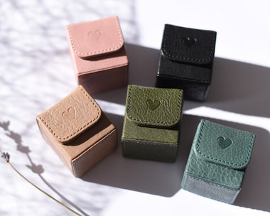 A collection of leather ring boxes in beige, black, green, and brown colors, each with a closed lid and a soft suede fabric pillow visible. A ring is placed on one of the open boxes, and there is a small flower beside the boxes.