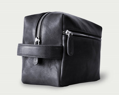 A zippered toiletry pouch made of black leather with a side handle and metal feet.