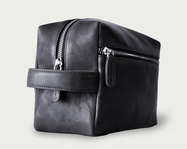 A zippered toiletry pouch made of black leather with a side handle and metal feet.