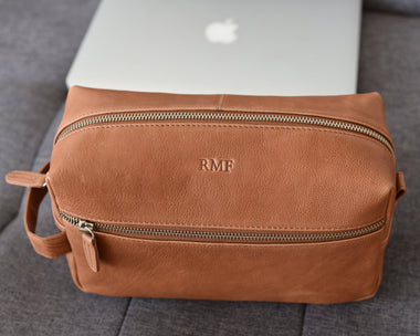 Light brown full grain leather men's toiletry bag with a hot stamped monogram on a grey couch.