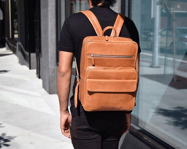 A person wearing a brown leather laptop backpack with a front zipper pocket, adjustable shoulder straps, and a magnetic flap closure. The backpack is shown from the front view on a city street.