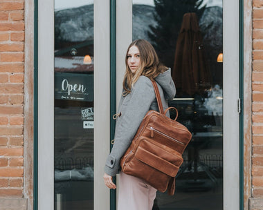 A person standing in front of a store window, wearing a gray jacket and carrying a brown leather laptop backpack with a zipper closure.