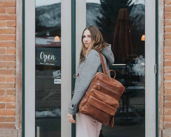 A person standing in front of a store window, wearing a gray jacket and carrying a brown leather laptop backpack with a zipper closure.