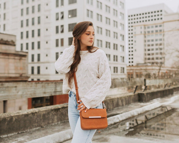 A model wearing a Caramel leather crossbody bag with a metal bar on the background of a large building