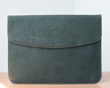 A dark green leather laptop sleeve with a front pocket and a flap closure, placed on a wooden surface.