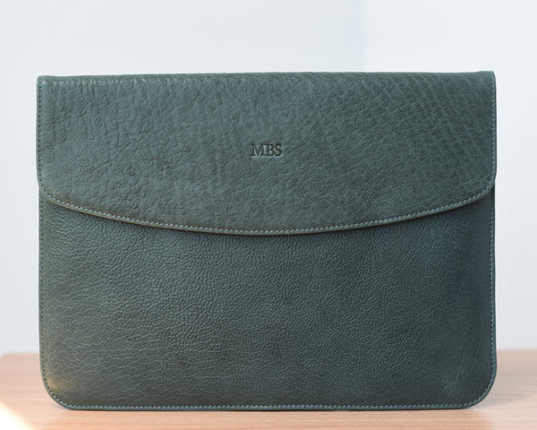 A dark green leather laptop sleeve with a front pocket and a flap closure, placed on a wooden surface.