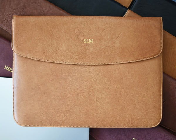 A brown leather laptop sleeve with a flap closure, positioned on top of other similar items. The sleeve features a card holder on the front and a full-length slip-in back pocket.