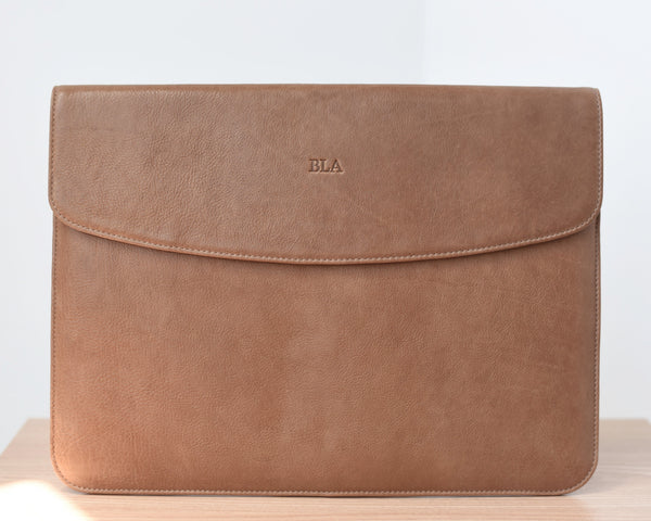 A light brown leather laptop sleeve with a front pocket and a flap closure, placed on a wooden surface.
