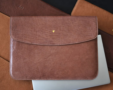A brown leather laptop sleeve with a front card holder and a back pocket, displayed on top of other brown leather items.