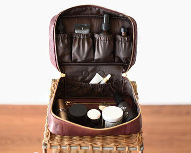Wine color leather cosmetic bag with toiletries inside shown open wide on a woven chair.