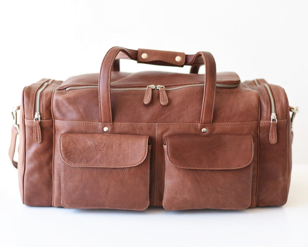 A large brown leather travel duffel bag with a shoulder strap, zippered compartments, and metal feet.